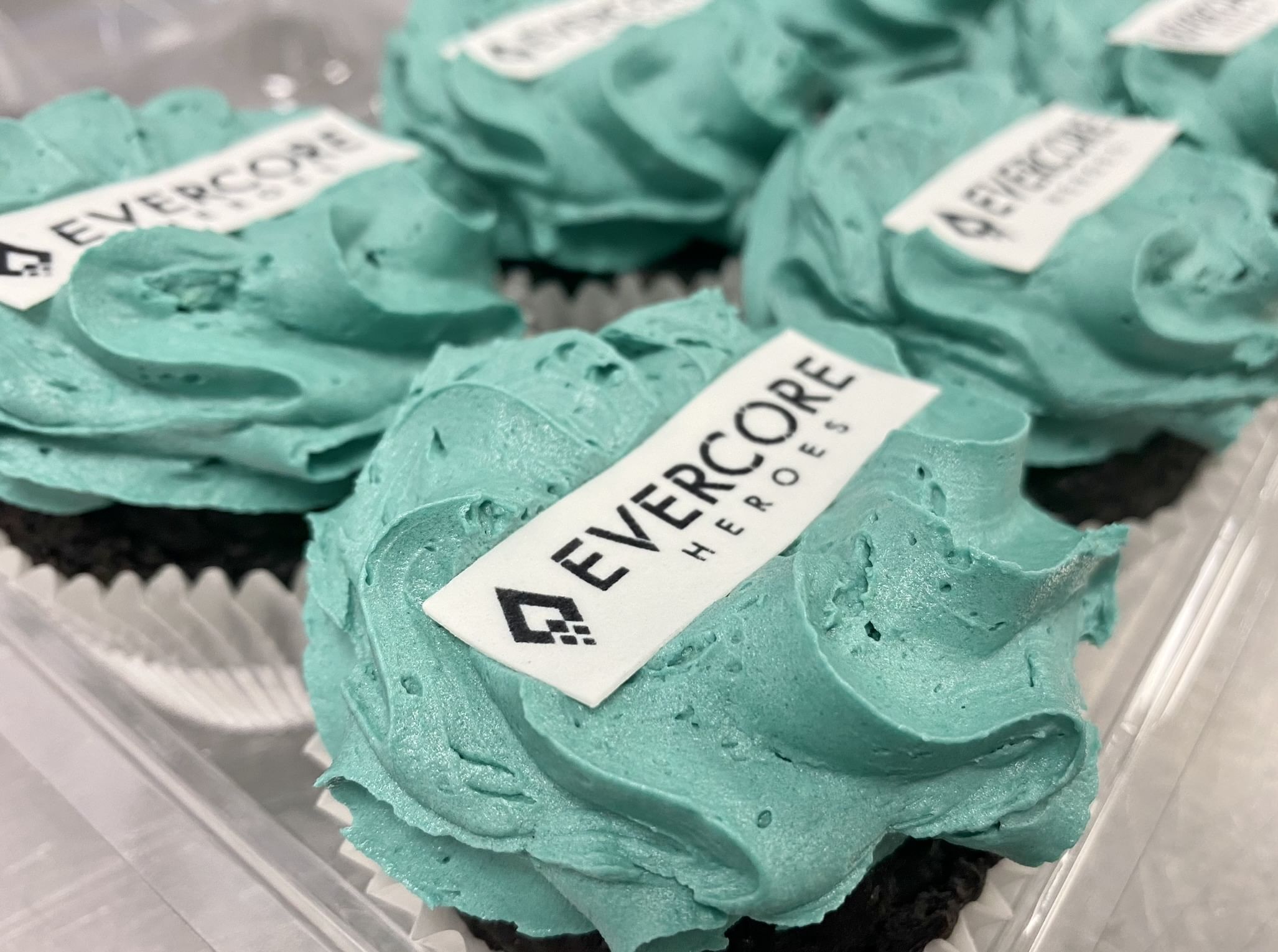 Close-up of custom-printed chocolate cupcakes with teal frosting and white edible logos that read 'EVERCORE HEROES.' These personalised cupcakes are arranged beautifully in a clear plastic container.